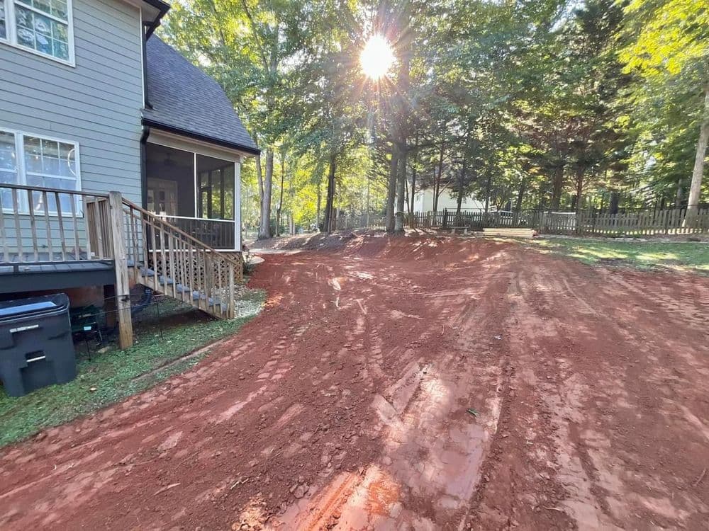 Sunlight filters through trees over a freshly tilled red dirt landscape beside a house.