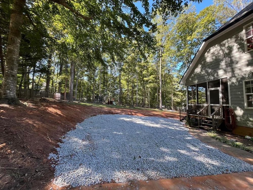 Gravel driveway installation beside a house in a wooded area. Clear blue sky overhead.