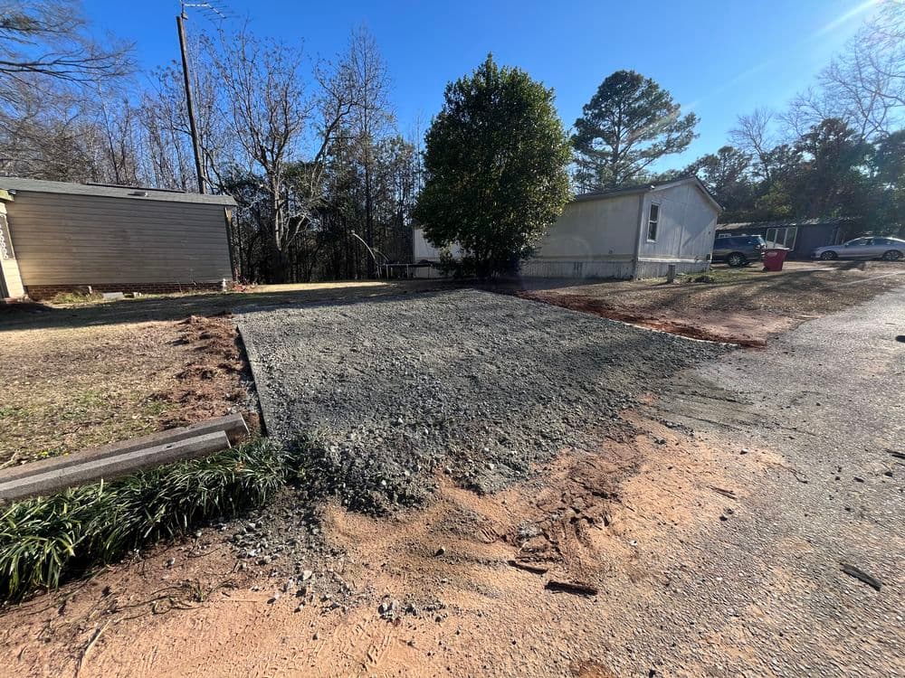 Gravel driveway prepared near a mobile home with trees and clear blue sky in the background.