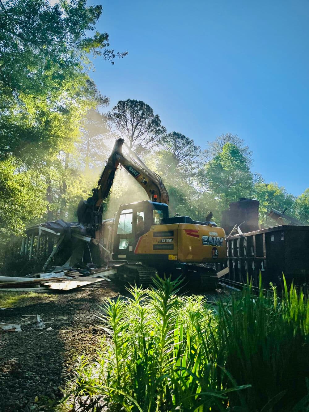 Excavator demolishing a structure in a sunny forested area with lush greenery.