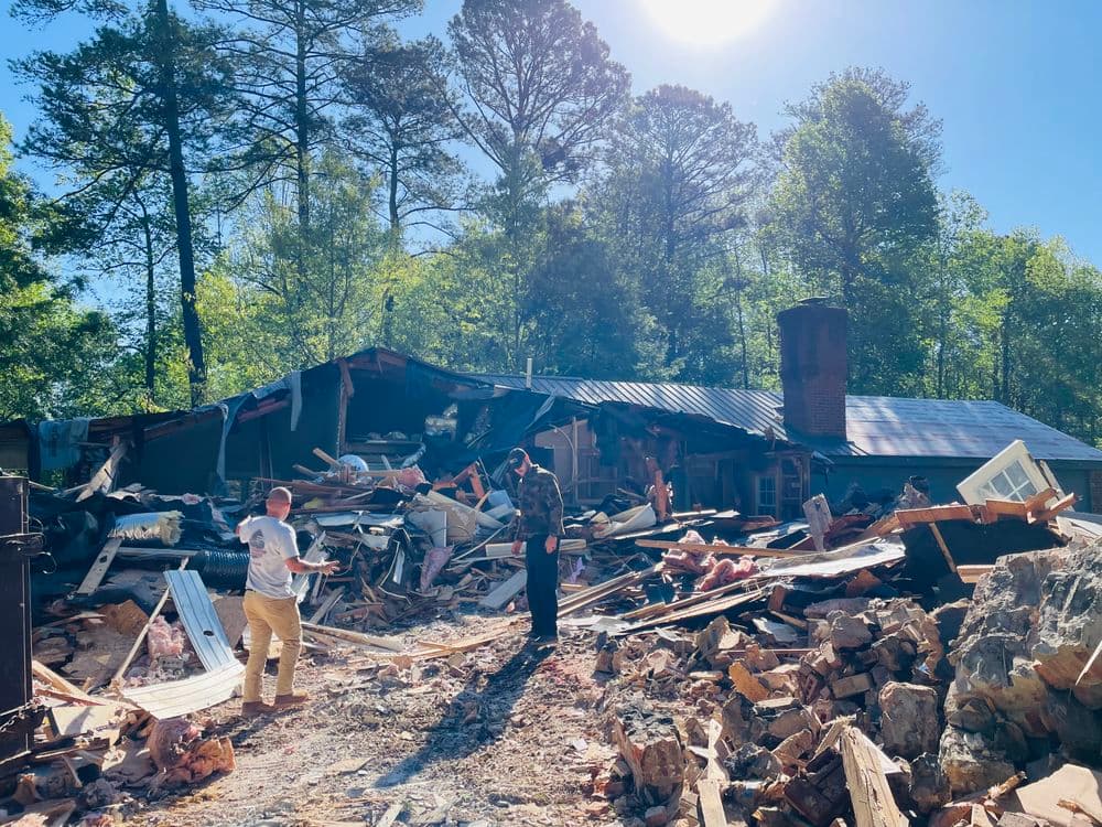 Collapsed house with two men assessing the damage in a wooded area.