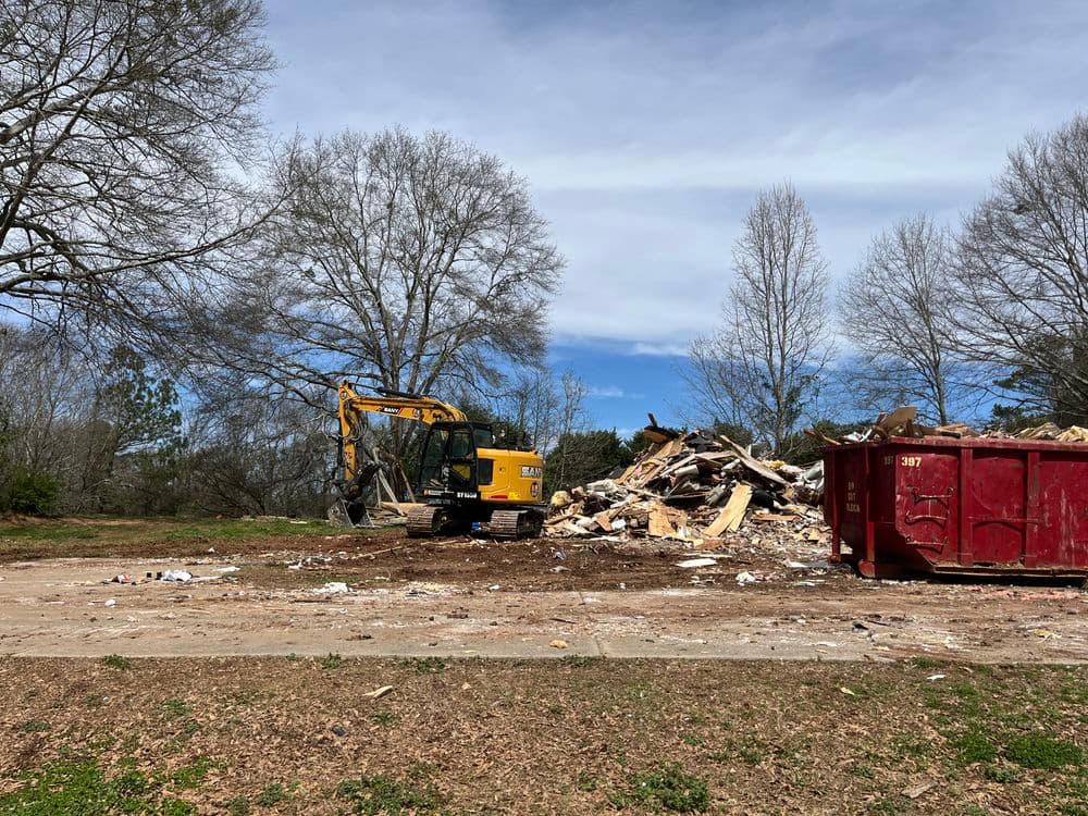 Construction site with excavator and dumpster amid debris under a cloudy sky.