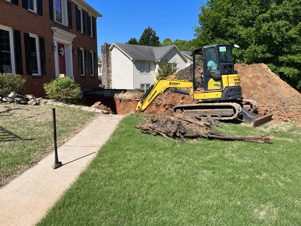 Excavator digging near residential home with dirt mound and trees on lawn.