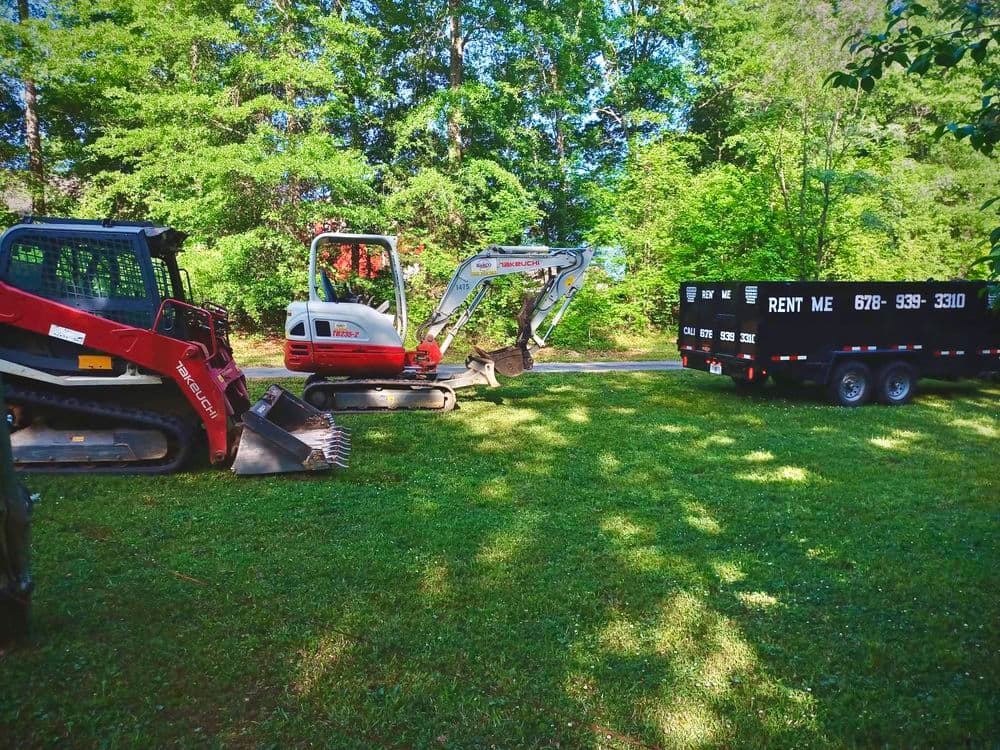 Excavation equipment and rental trailer on green grass in a wooded area.