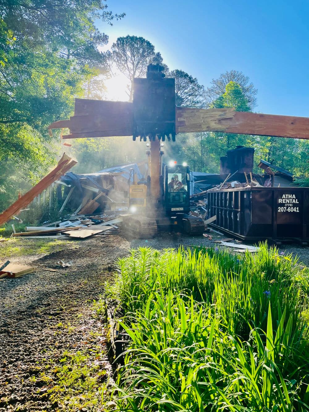Construction equipment demolishing a building surrounded by greenery and sunlight.