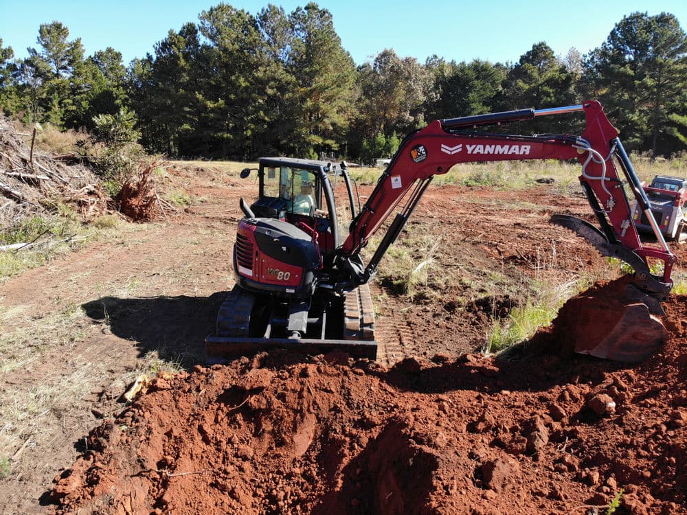 Yanmar excavator digging in a cleared area surrounded by trees and dirt mounds.