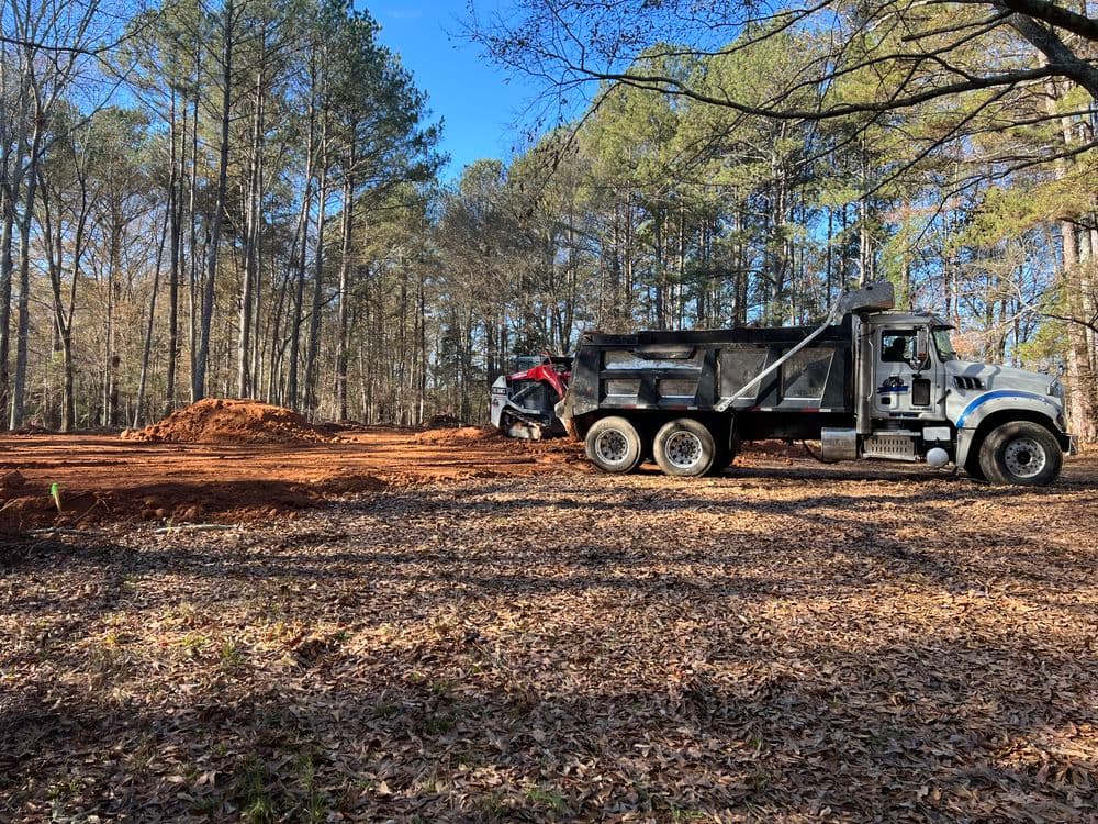 Construction site with a dump truck unloading soil in a wooded area.