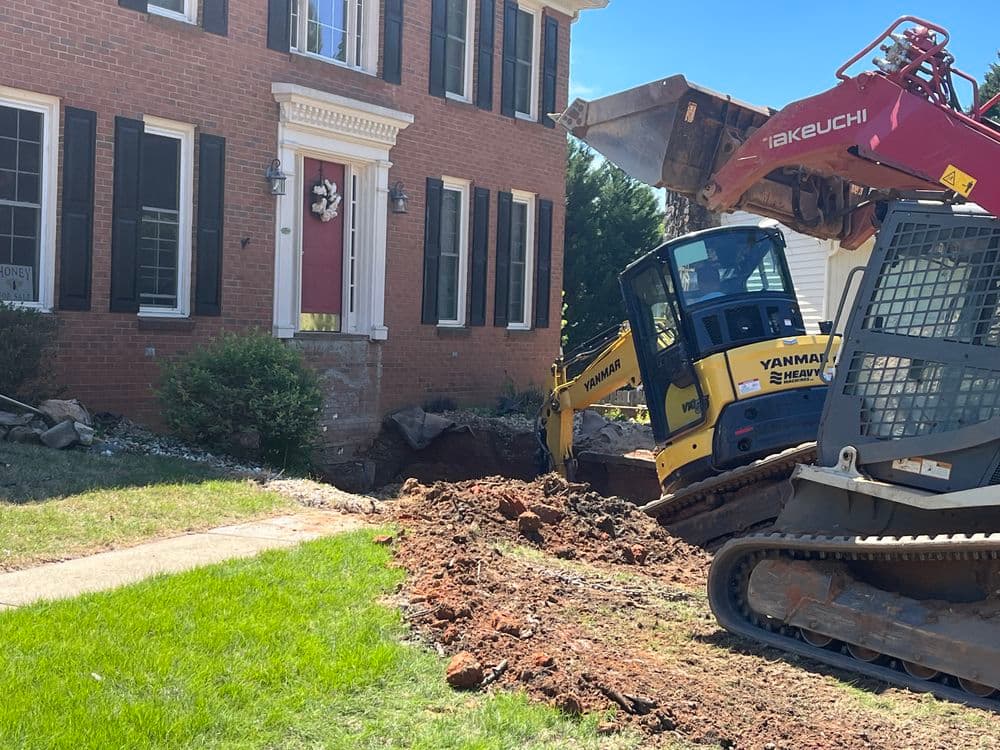 Excavation equipment digging near a brick house with landscaping in progress.