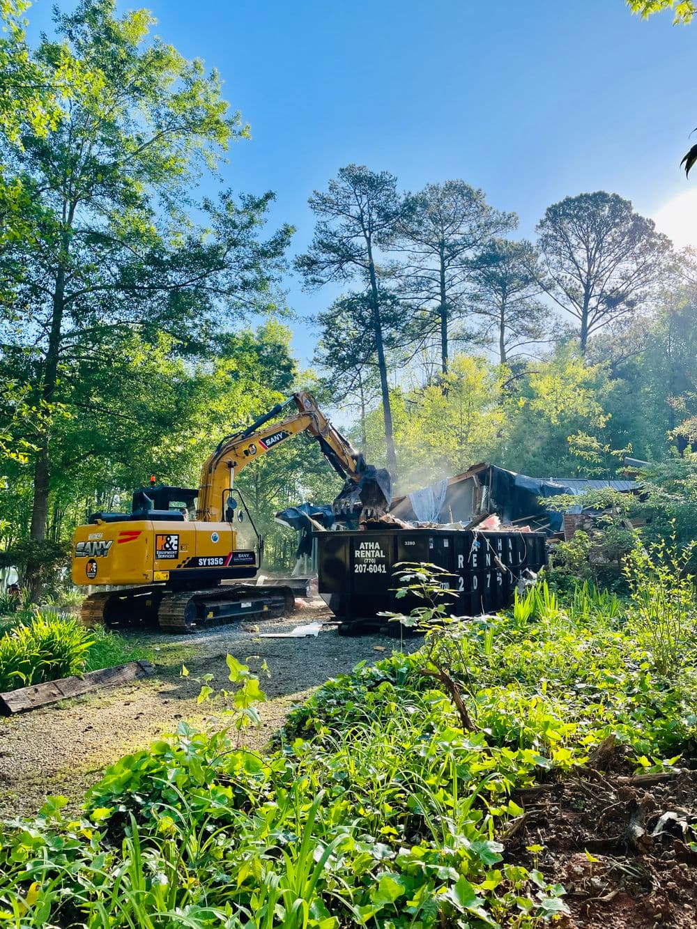 Excavator demolishing a structure near a dumpster in a wooded area on a sunny day.