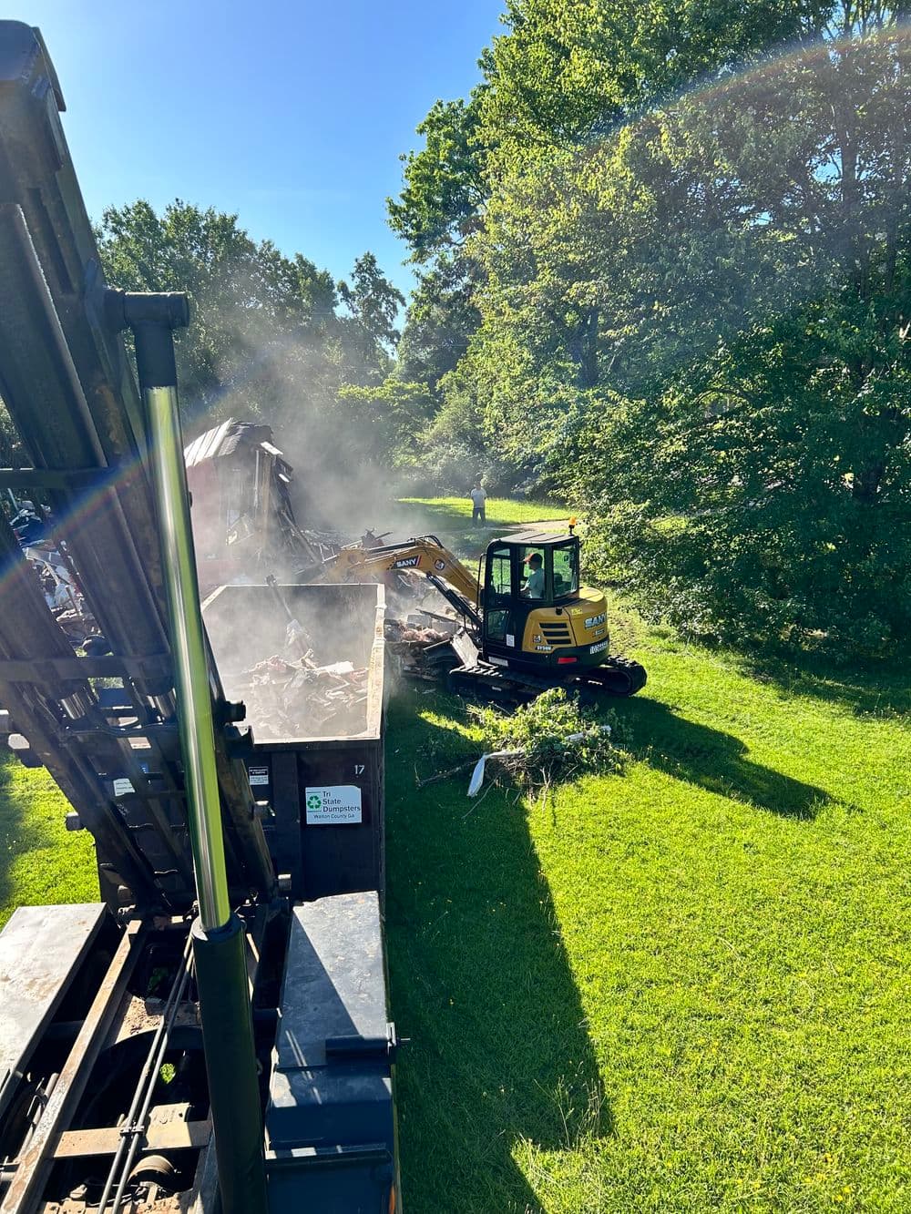 Excavator loading debris into a dumpster during demolition on a sunny day.