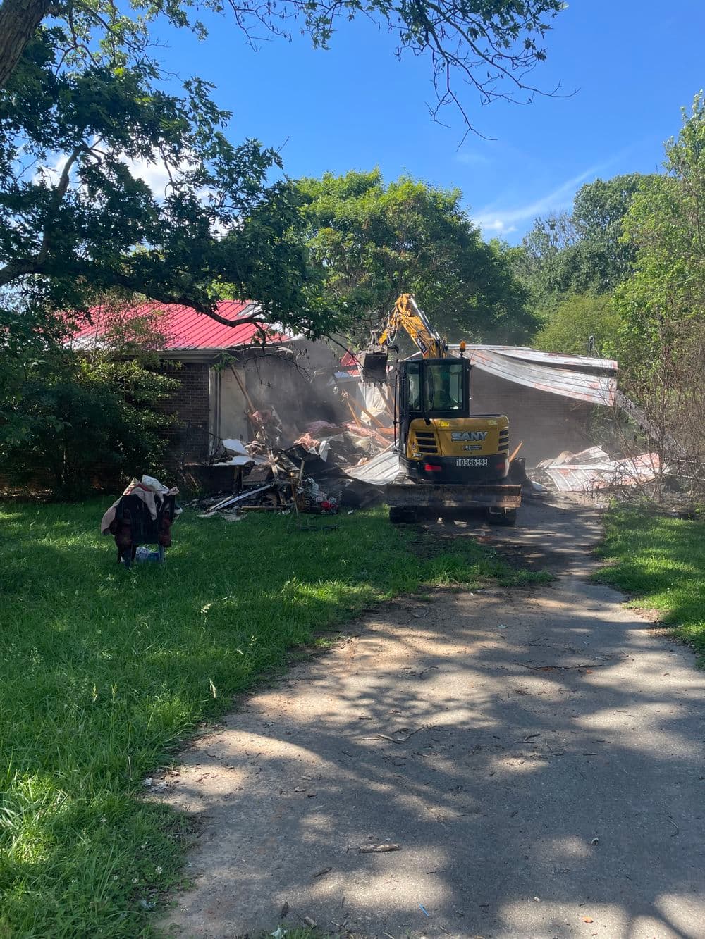 Excavator demolishing a house with debris in a grassy area under blue sky.
