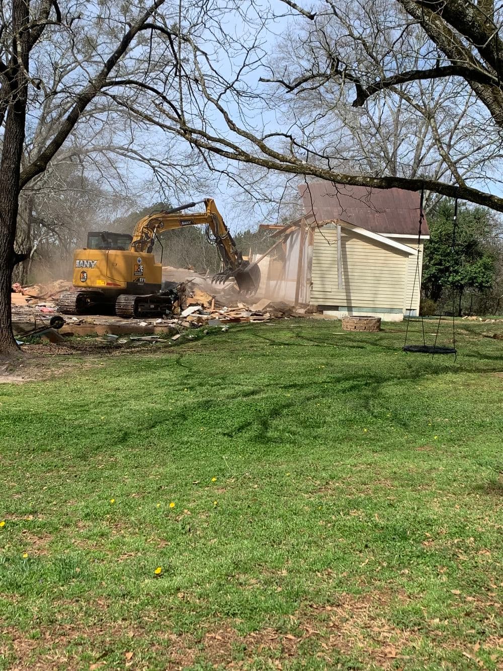 Excavator demolishing a house in a grassy yard with trees and dust in the air.