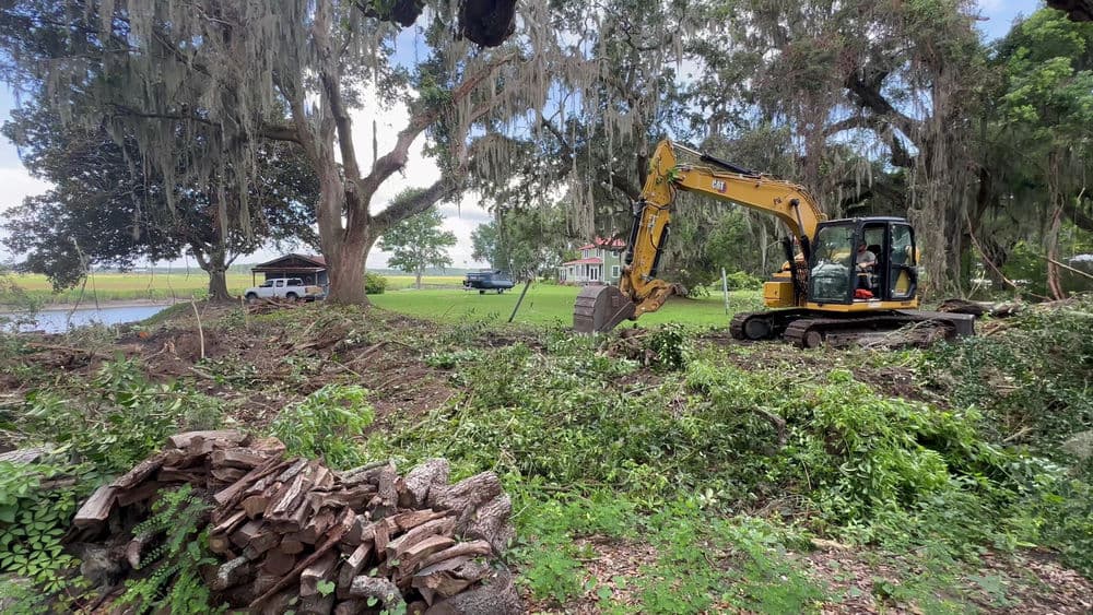 Excavator clearing debris in a lush, tree-filled yard with a farmhouse in the background.