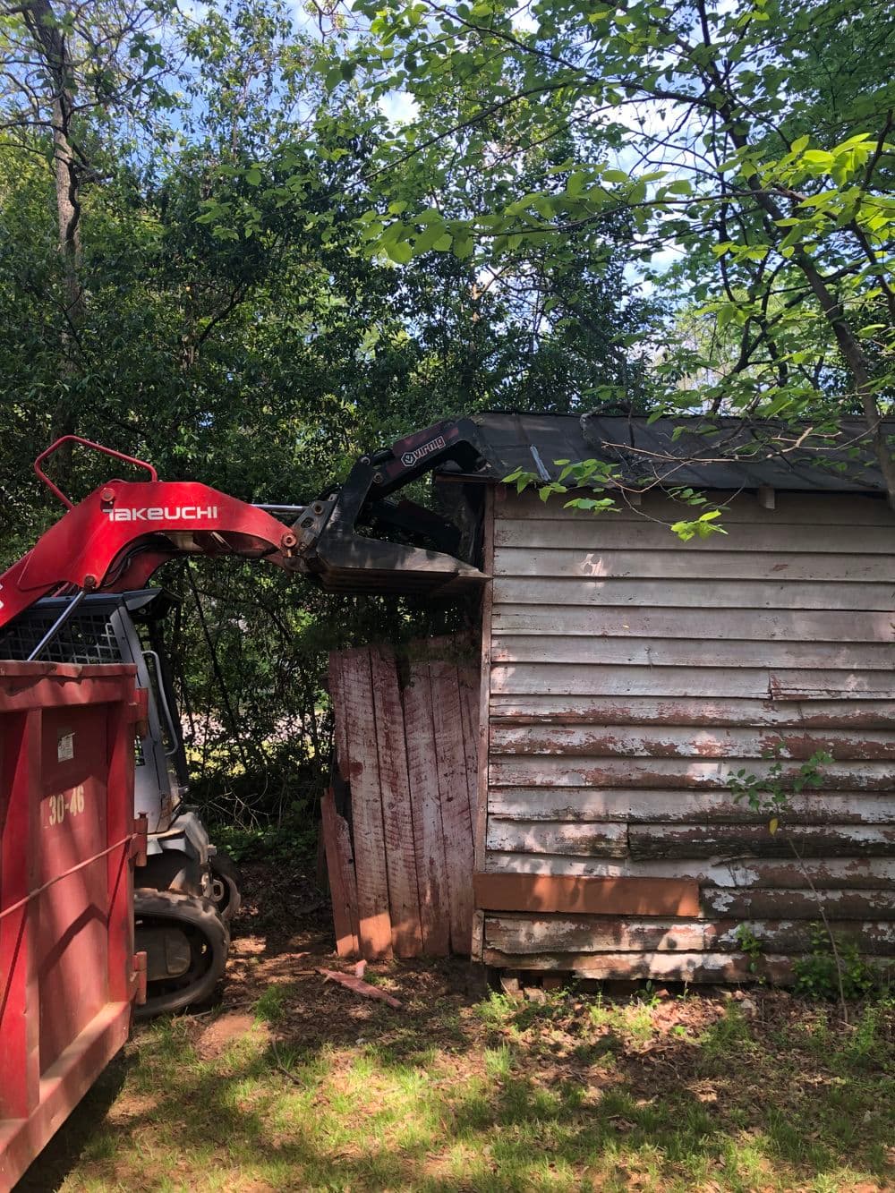 Skid steer loader removing debris from an old wooden shed in a wooded area.