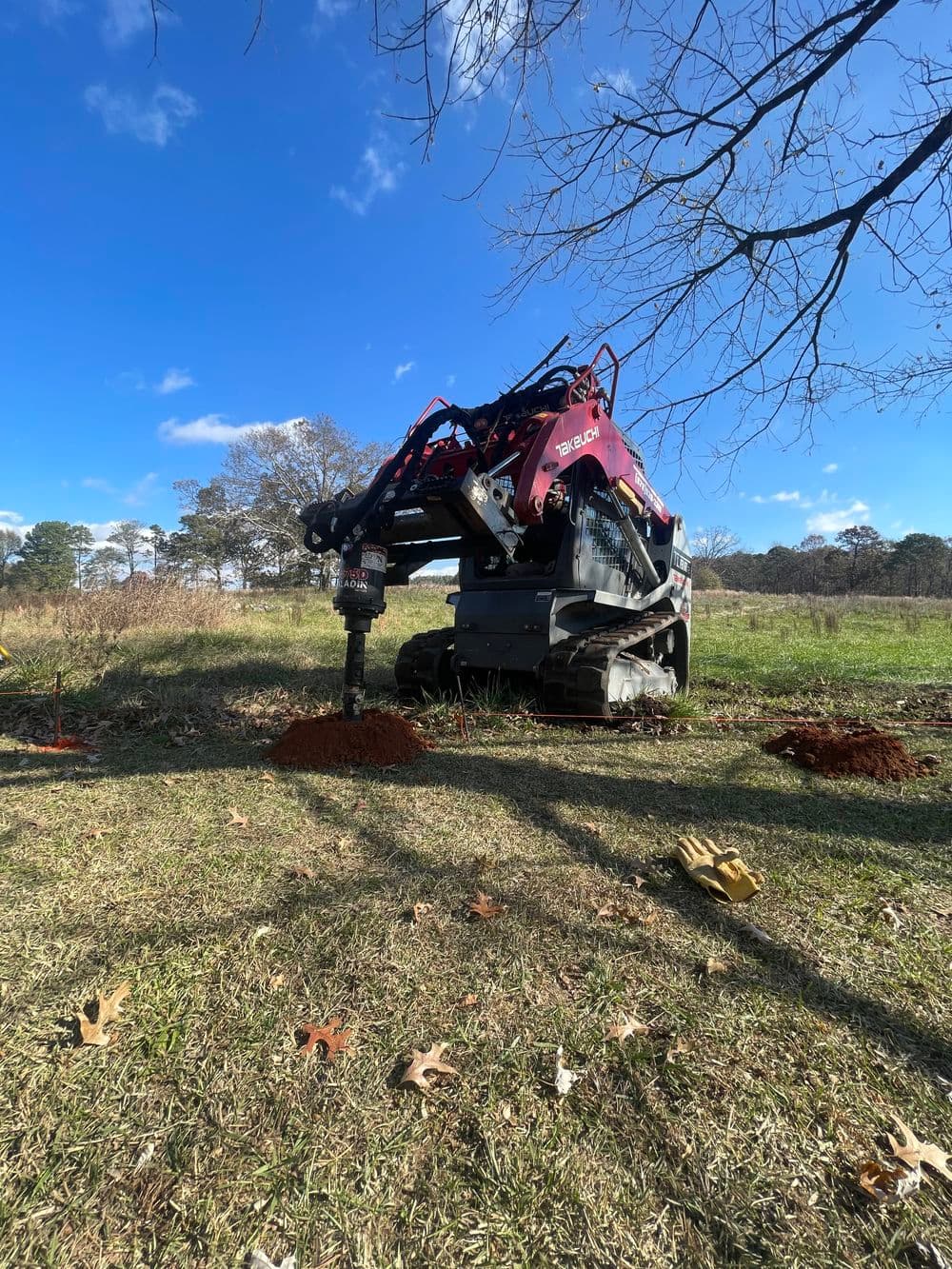 Compact track excavator drilling into the ground on a sunny day in a rural setting.