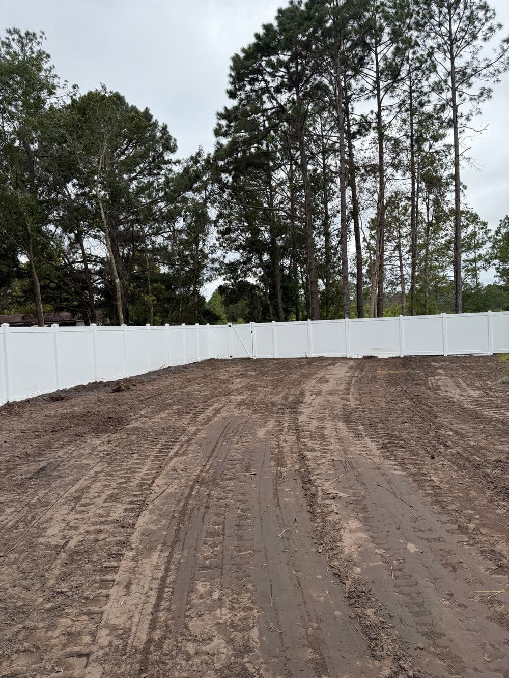 White vinyl fence surrounding a cleared dirt area, bordered by tall trees.